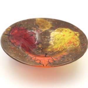 A glass circular bowl with yellow, red nad orange maple leaves and a brown and amber background accented with bronze and black specks