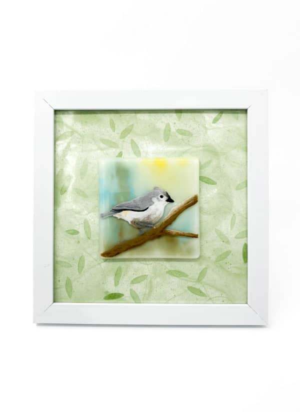 A grey and white tufted titmouse bird perched on a branch whith a pale yellow, green and blue background. The bird is an enamel painting on a glass tile that is 4x4". It is framed in a 8x8 frame with a pale green leaf patterned mat.