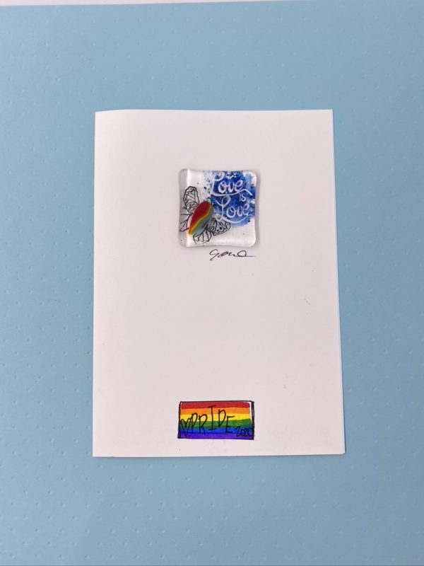 5x8 greeting card with attached 1.5" square fused glass art featuring a delicate filigree style butterfly with a rainbow striped body and the words "Love is Love" painted above the butterfly. On the bottom center of the card face is a rainbow flag with "Pride 2020" printed on top