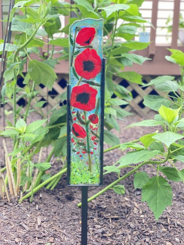 five varied size poppy blossoms on a blue and green background in a black frame which is also the stake for this garden art