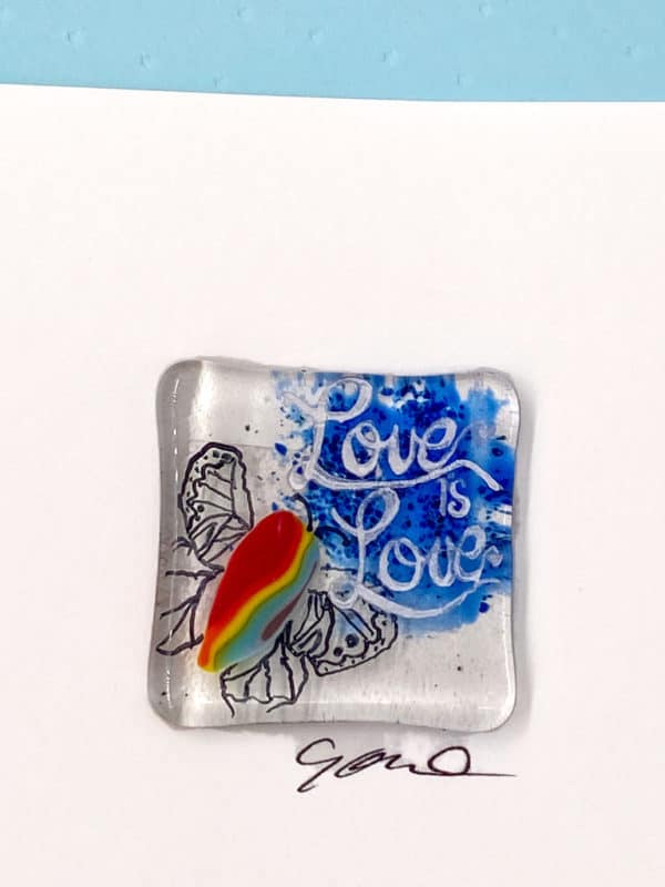 close up photo of a 1/5" square filigree winged butterfly with a rainbow striped body. The words "Love is Love" are painted above the butterfly over a blue background