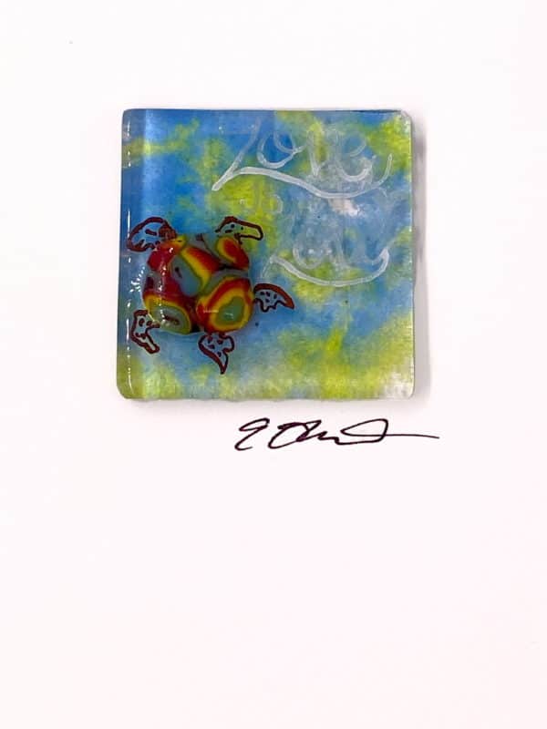 a rainbow shell fused glass sea turtle is swimming on a green and blue background, the words "Love is Love" are painted in white. This is a closeup photo of a 2" square piece of fused glass art
