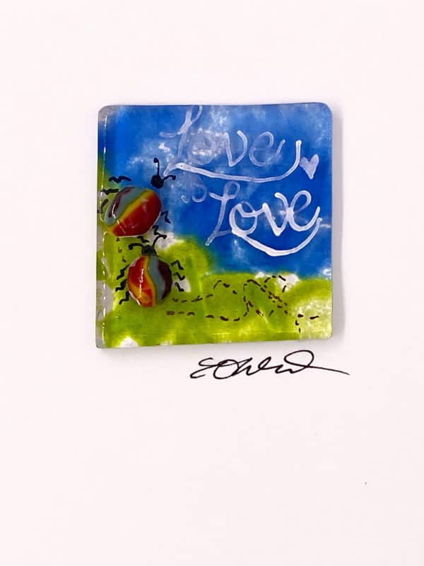 close up of a 2x2 miniature fused glass art with two rainbow body bugs on a green and blue background with the words Love is Love in the right hand corner