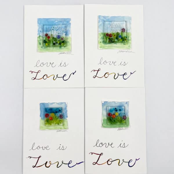 four greeting cards that have a 1.5 inch square tiny rainbow flower garden on a watercolor mat background. The text of the card says "Love is Love" in hand drawn modern calligraphy style and the second "Love" is large and highlighted with the Pride rainbow colors