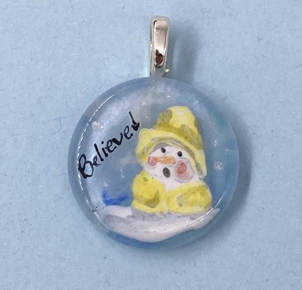1" glass circle pendant with painting of a snow man dressed. in a yellow rain slicker next to the word "Believe"