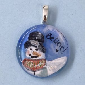 1" circle glass pendant with snow person in a top had and red/green checked vest with the word "Believe" next to it