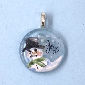 1" enamel painted glass pendant with a snow person in a top hat and green scarf and the word "Joy" written in the sky next to it
