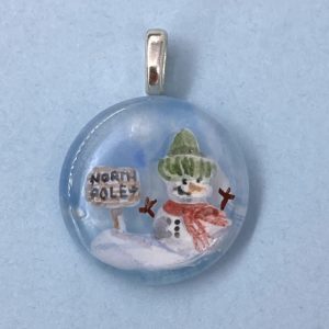 1" circle pendant with hand painted snow man wearing a green winter hat and red scarf next to a wooden sign that says "North Pole"