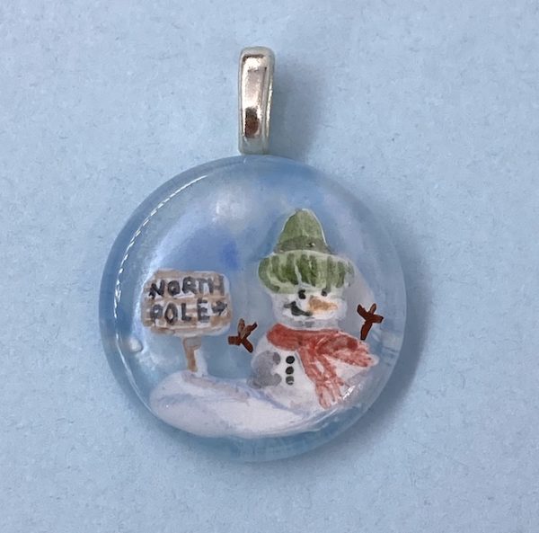 1" circle pendant with hand painted snow man wearing a green winter hat and red scarf next to a wooden sign that says "North Pole"