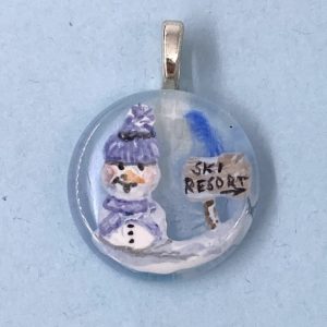 1" glass circle pendant with enamel painted snow man in purple knit cap and scarf next to a wooden sign that says "Ski Resort"