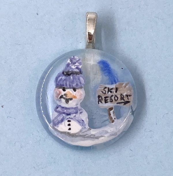 1" glass circle pendant with enamel painted snow man in purple knit cap and scarf next to a wooden sign that says "Ski Resort"