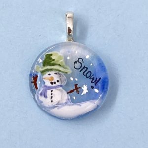 1" circle glass pendant with enamel painting of a snow man wearing a green top hat and purple scarf with the word "Snow" in the sky next to it