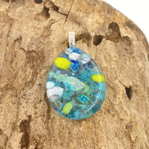 Photo is of a multicolored tear drop shaped pendant with a silver bail. There are multiple colors throughout the pendant which is made of many pieces of glass melted together, it is primarily teal with bits of white, yellow-green, cobalt and a very shiny focal piece near the center which is green/pink