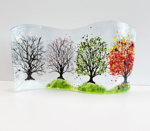 A glass art piece that features a tree in four seasons, the piece has a wavy top edge and is itself wavy and stands on its edge. The winter tree has a tiny red cardinal and sits in snow, the spring tree has pink blossoms and sits on green grass, the summer tree has green leaves and is on green grass and the autumn tree has yellow, orange and red glass leaves and sits on green grass with scattered colored leaves sitting on it