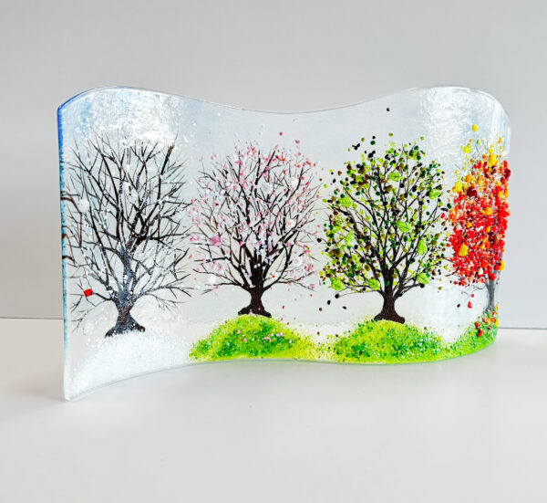 A glass art piece that features a tree in four seasons, the piece has a wavy top edge and is itself wavy and stands on its edge. The winter tree has a tiny red cardinal and sits in snow, the spring tree has pink blossoms and sits on green grass, the summer tree has green leaves and is on green grass and the autumn tree has yellow, orange and red glass leaves and sits on green grass with scattered colored leaves sitting on it