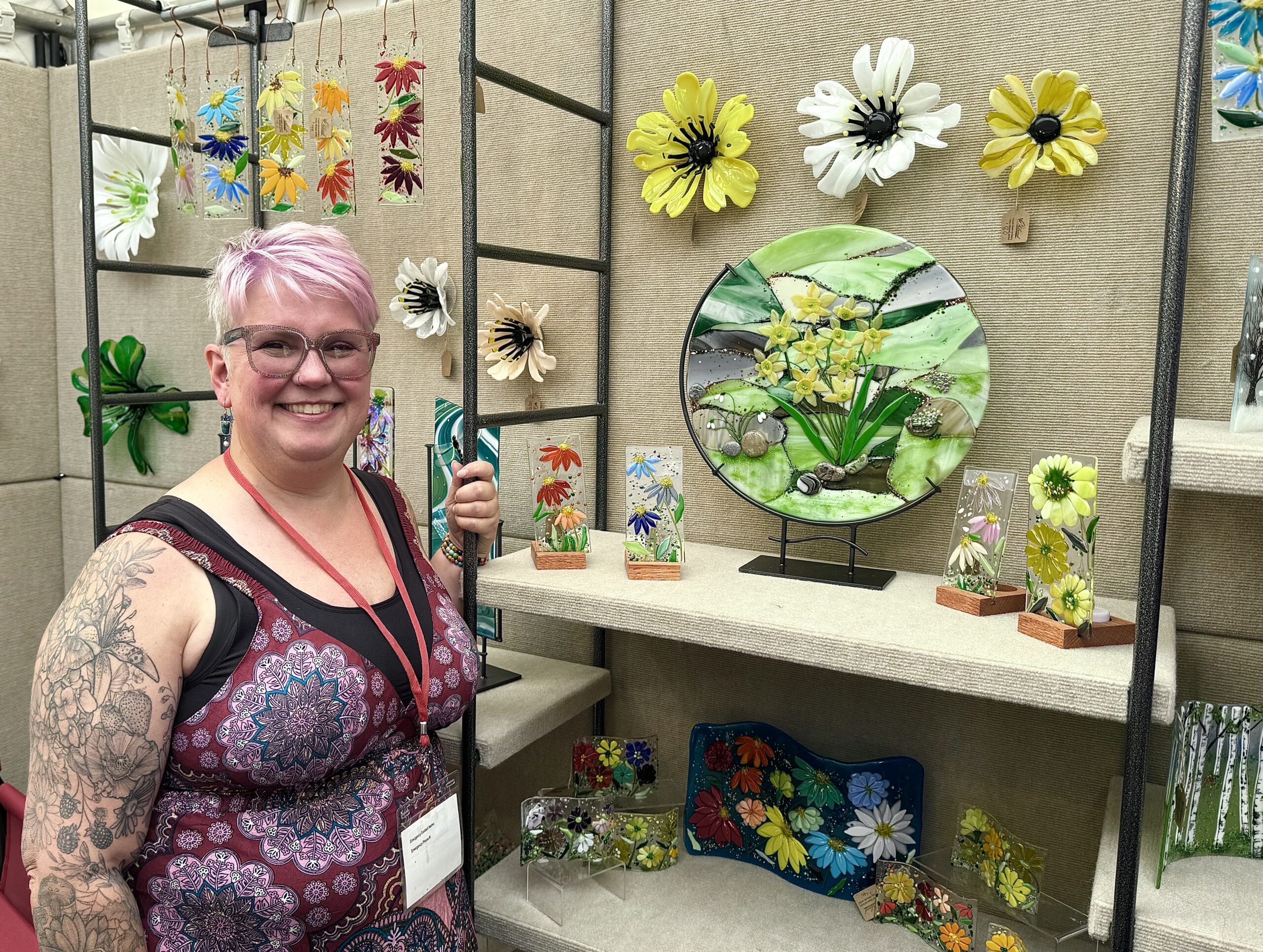 184DE5DB-7A3B-4B15-B5BB-B71291A229C4 a woman with gray & purple short hair is standing in front of an art fair booth display that has many works of fused glass art