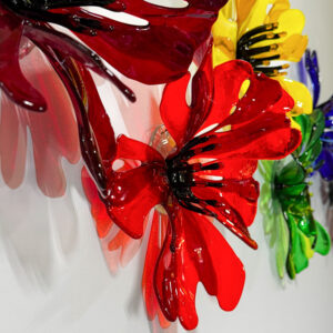 A collection of brightly colored 3D glass flowers loosely poppy-like in red, orange, yellow green and blue. The flowers are hanging on a wall and the light travels through the glass and casts colorful shadows on the wall