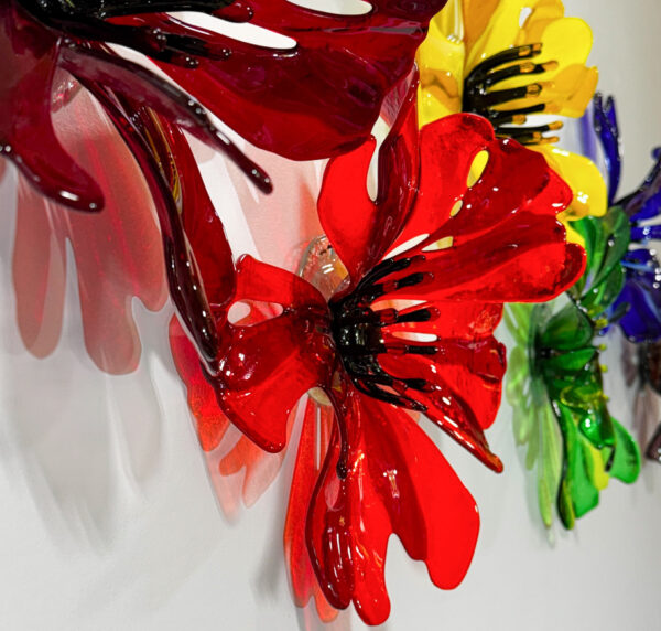 A collection of brightly colored 3D glass flowers loosely poppy-like in red, orange, yellow green and blue. The flowers are hanging on a wall and the light travels through the glass and casts colorful shadows on the wall