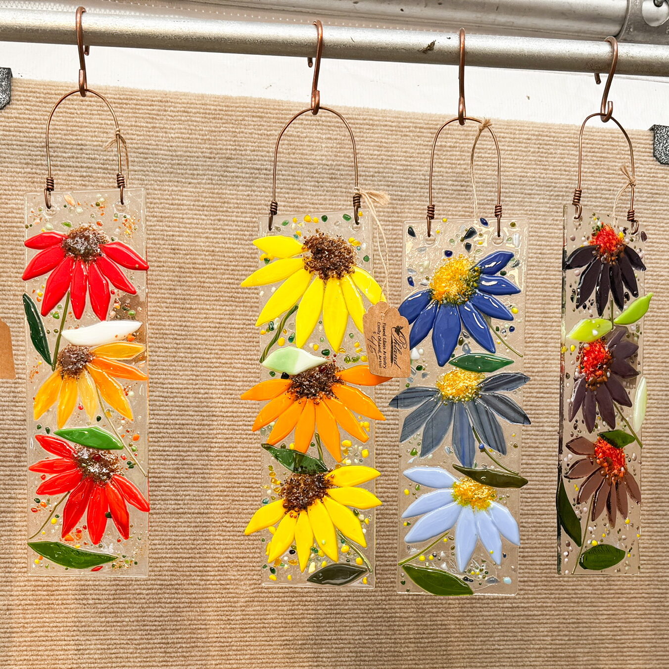 image of 6 hanging floral glass art pieces, each piece is topped with a copper wire loop for hanging and has 3 flowers vertically. The colors in each piece vary in hue. Each individual glass piece is 9x3" and from left to right there are red poppies, orange daisy/cone flowers, yellow daisy/cone flower, blue cone flowers, purple cone flowers and pink/ivory cone flowers