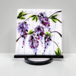 Custom Wisteria Art Panel image is a glass art panel of 5 wisteria flower clusters on a white and coordinating purple/blue/pink background. There are green leaves and brown stems and the flowers appear to have grown in from the sides of the piece